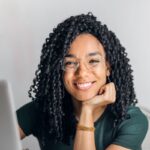 Joyful businesswoman with curly hair smiling at camera while using laptop indoors.